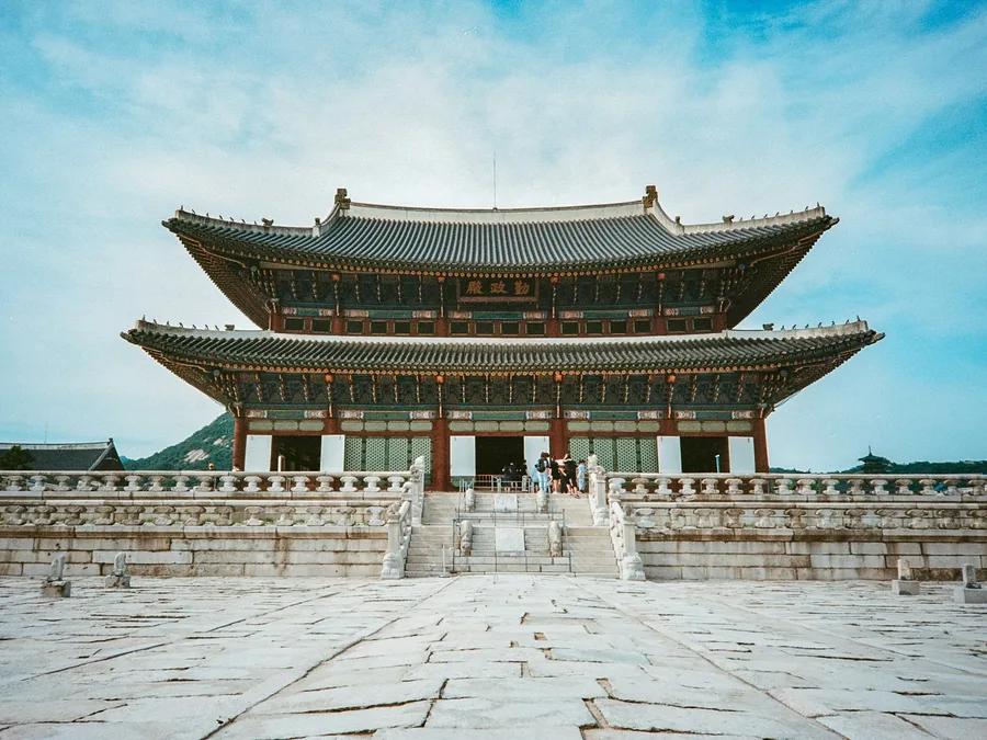 Foto Istana Gyeongbokgung di Seoul: aula kayu bertingkat dua beratap hijau di atas pelataran batu, di bawah langit biru.
