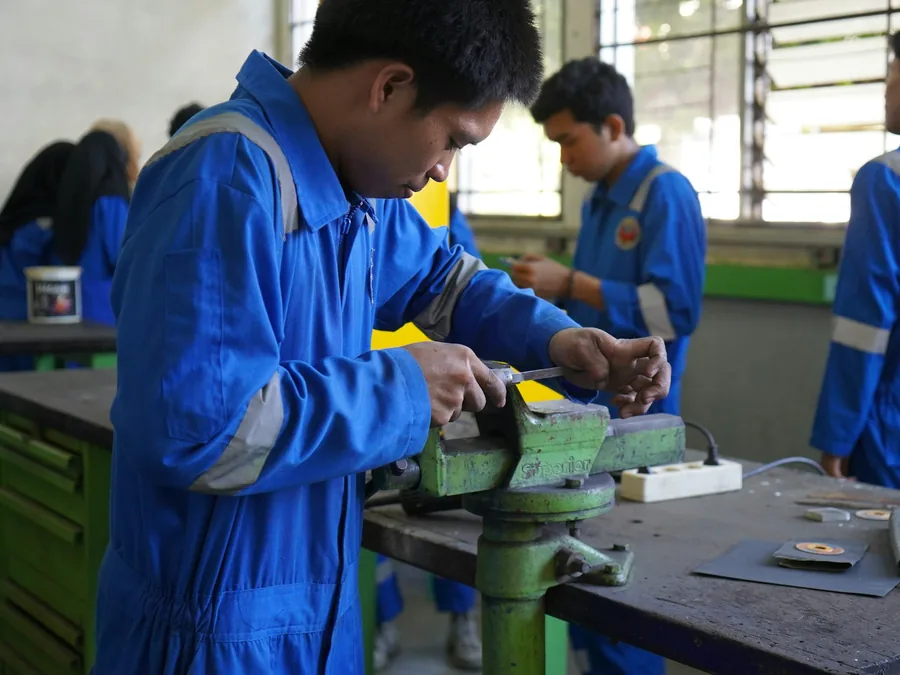 Foto siswa pelatihan kejuruan berbaju kerja biru mengerjakan benda logam di ragum, dengan siswa lain di bengkel praktik.