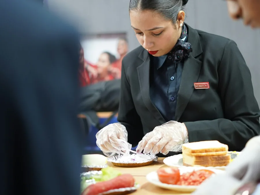 Foto peserta pelatihan kuliner berjas gelap dan bersarung tangan plastik menyiapkan roti lapis di meja dapur.